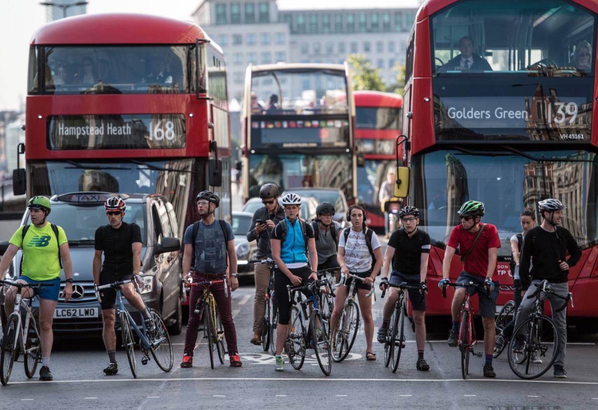 The image shows a crowd of white, male cyclists in front of a few cars and four busses.